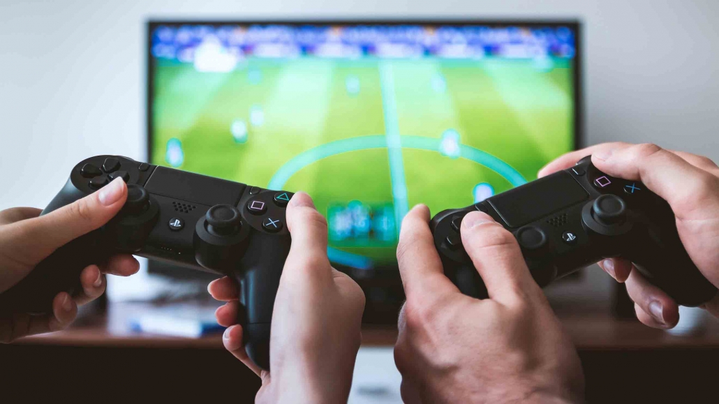 Auch beim Zocken kannst Du Energie sparen - wir haben die passenden Energiespar-Tipps für Dich zusammengefasst. Zwei Controller vor Fernseher, auf dem Fußballspiel gespielt wird.