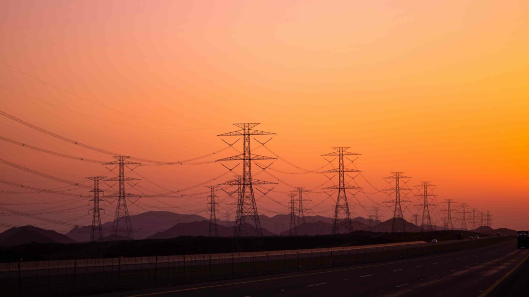 Hochspannungs-Strommasten vor einem orangefarbenen Himmel bei Sonnenuntergang, die Stromleitungen transportieren. Symbolisiert den Transport von Energie durch das Stromnetz in ländliche und städtische Gebiete.