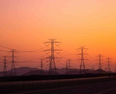 Hochspannungs-Strommasten vor einem orangefarbenen Himmel bei Sonnenuntergang, die Stromleitungen transportieren. Symbolisiert den Transport von Energie durch das Stromnetz in ländliche und städtische Gebiete.