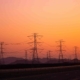 Hochspannungs-Strommasten vor einem orangefarbenen Himmel bei Sonnenuntergang, die Stromleitungen transportieren. Symbolisiert den Transport von Energie durch das Stromnetz in ländliche und städtische Gebiete.
