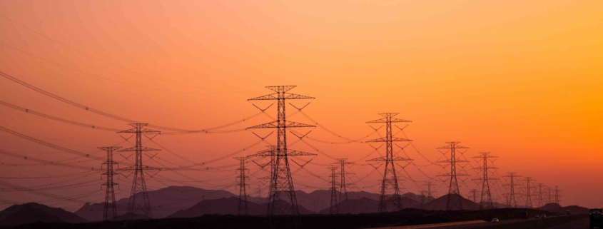 Hochspannungs-Strommasten vor einem orangefarbenen Himmel bei Sonnenuntergang, die Stromleitungen transportieren. Symbolisiert den Transport von Energie durch das Stromnetz in ländliche und städtische Gebiete.